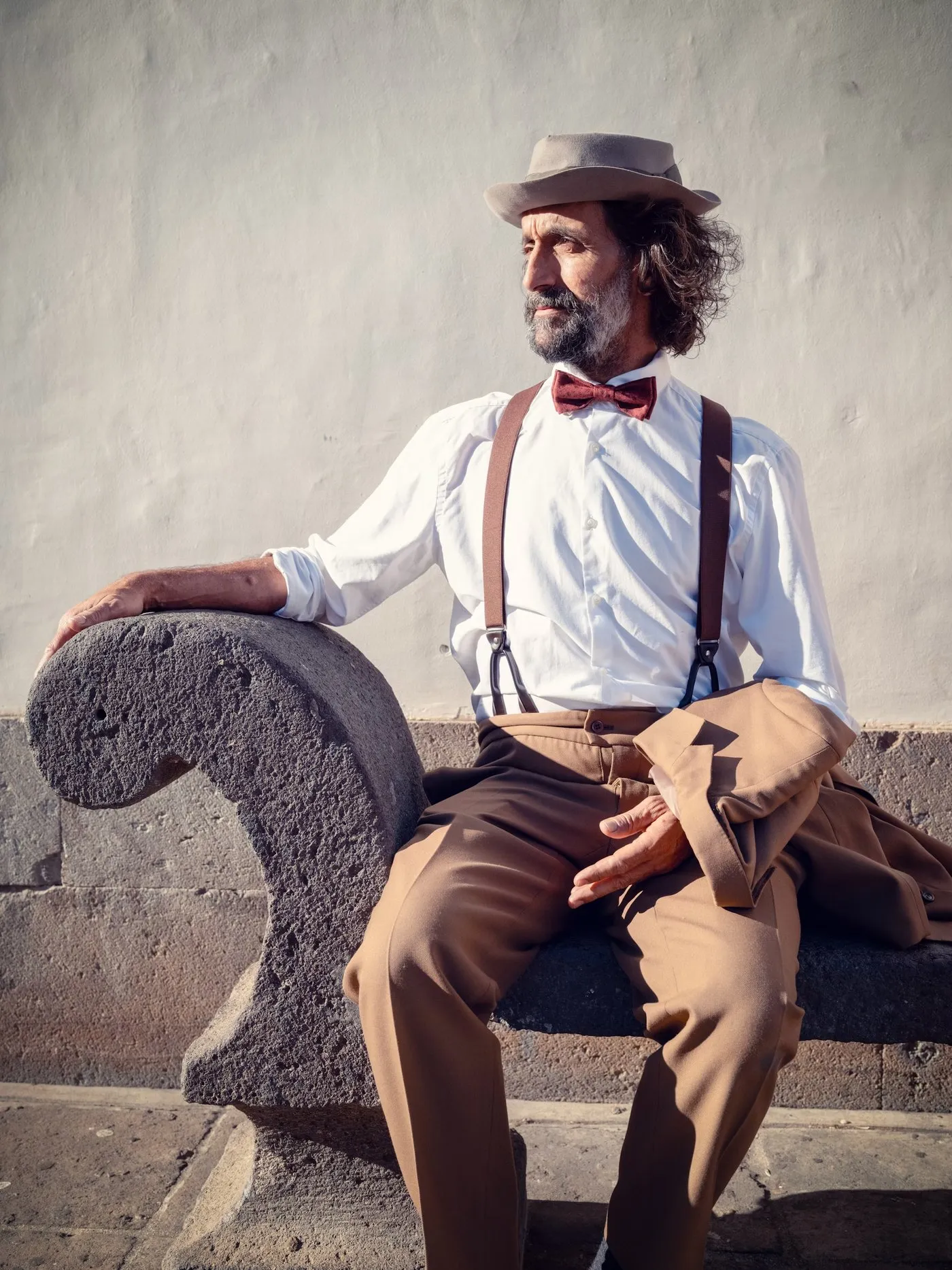 Robert Fox in a hat and bow tie, sitting on a stone wall